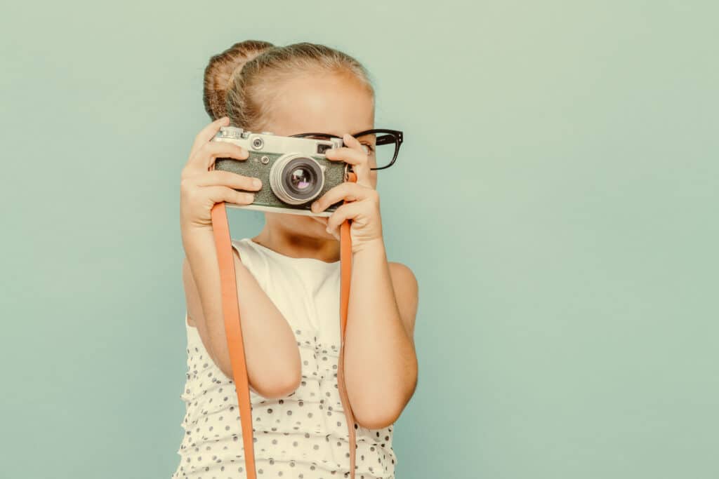 Stock photo of a girl wearing thick black glasses and holding a camera up to her eye. She has blonde hair tied back into a bun.