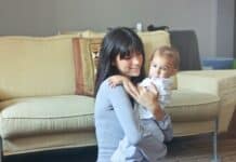 Stock photo of a woman with straight brown hair and bangs holding an infant in her arms in a living room. She is smiling at the baby, and the baby is looking at the camera.