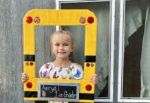 DIY Back-to-School Photo Frame Photo of a young girl holding a homemade photo frame and smiling at the camera. She is wearing her blonde hair in a ponytail and is wearing a white dress with colorful butterflies on it.