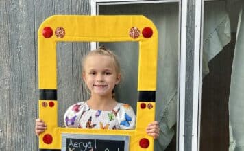 Photo of a young girl holding a homemade photo frame and smiling at the camera. She is wearing her blonde hair in a ponytail and is wearing a white dress with colorful butterflies on it.