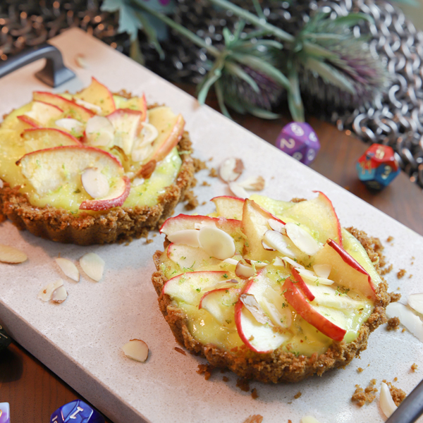 Photo of two small tart pastries topped with thin apple slices on a wooden rectangular platter. Sliced almonds are scattered around the tarts.