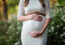 Stock photo of a pregnant woman holding her stomach with both hands and standing outside. She is wearing a white dress with a thin brown leather belt around her waist.