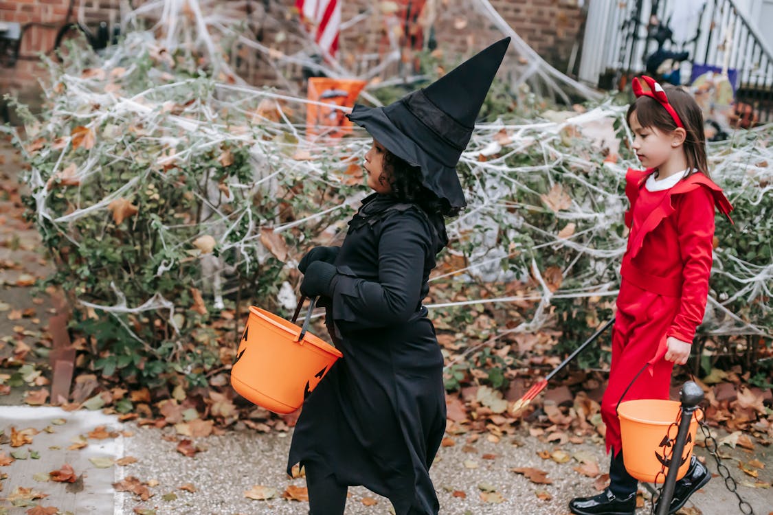 Stock photo of two children dressed up in costume holding small orange buckets for trick-or-treating. The one on the left is wearing all black and a pointed witch's hat and the other is dressed in red with a red devil's horn headband.
