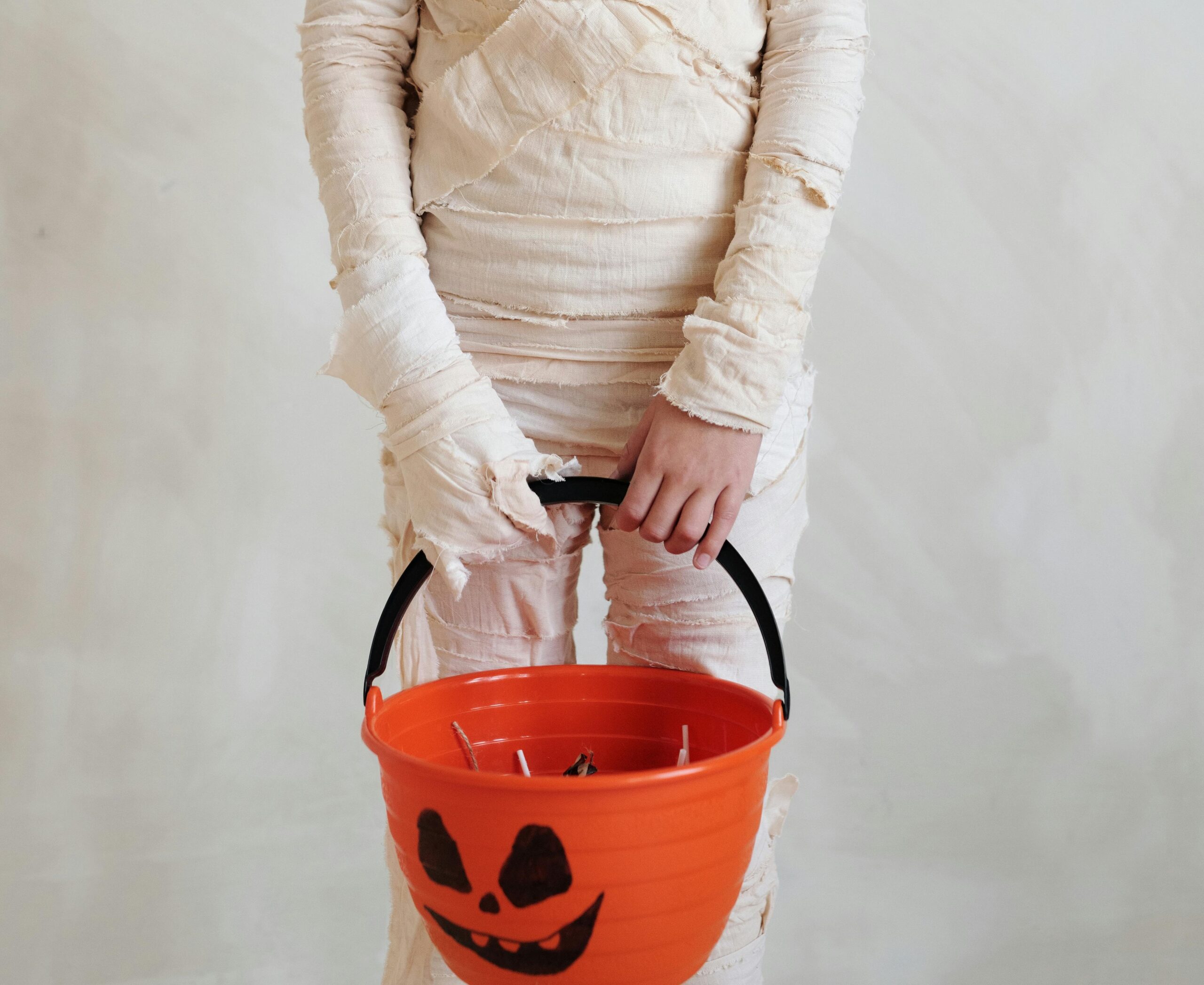 Stock photo of a child in a mummy costume holding an orange jack-o-lantern bucket.
