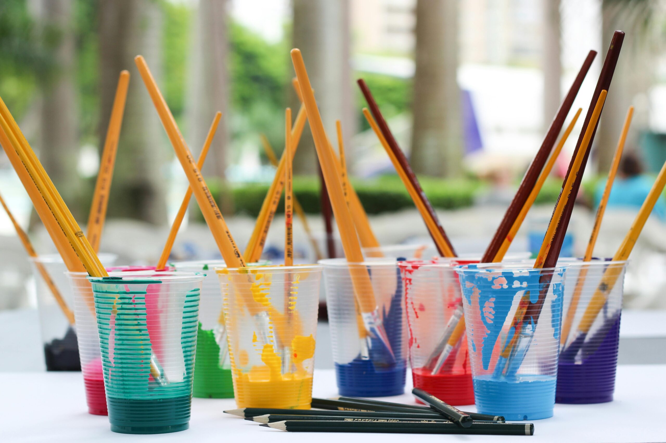Stock photo of a half dozen plastic cups holding different colored water and a paint-covered paintbrush on an outdoor table.