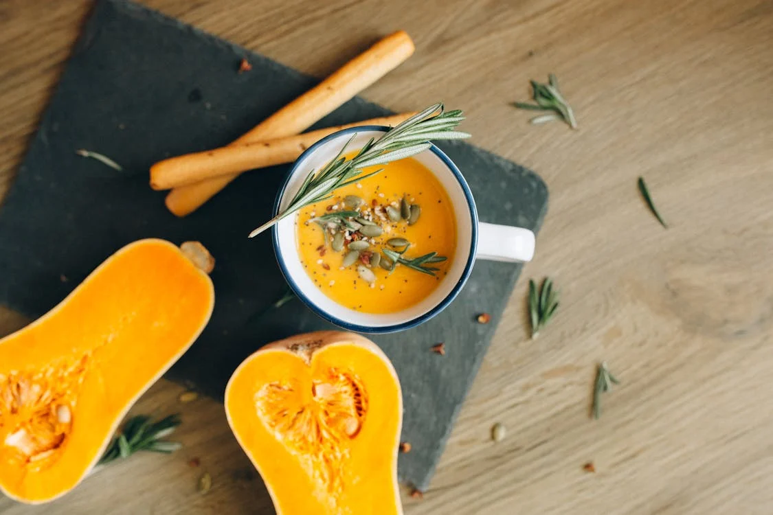 Stock photo of orange pumpkin soup in a mug topped with green herbs. On the table is a squash cut into halves.