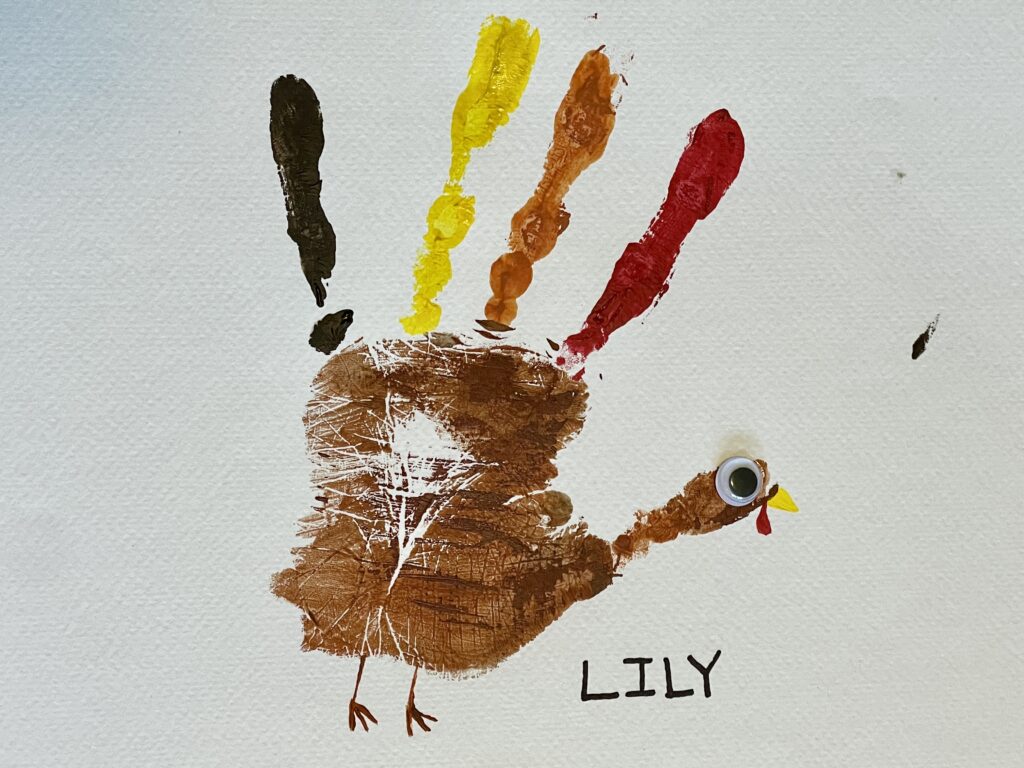 Photo of a child's painted handprint in the colors of a turkey. The palm is painted brown and the fingers are red, orange, yellow and darker brown. The turkey has a googly eye and a red gobble under its mouth. Writing in pen reads "Lily" next to the turkey handprint.