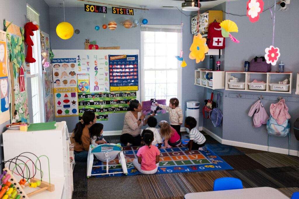 Photo of the colorful interior of a daycare with toddlers sitting and playing on the floor. The walls are painted periwinkle and there are hanging mobiles and toys around the room.