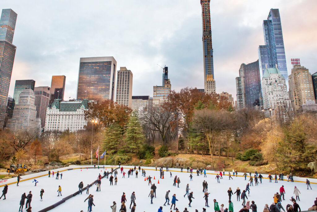Wide shot of a large outdoor ice skating rink with dozens of skaters dressed in winter gear. New York City skyscrapers line the background.