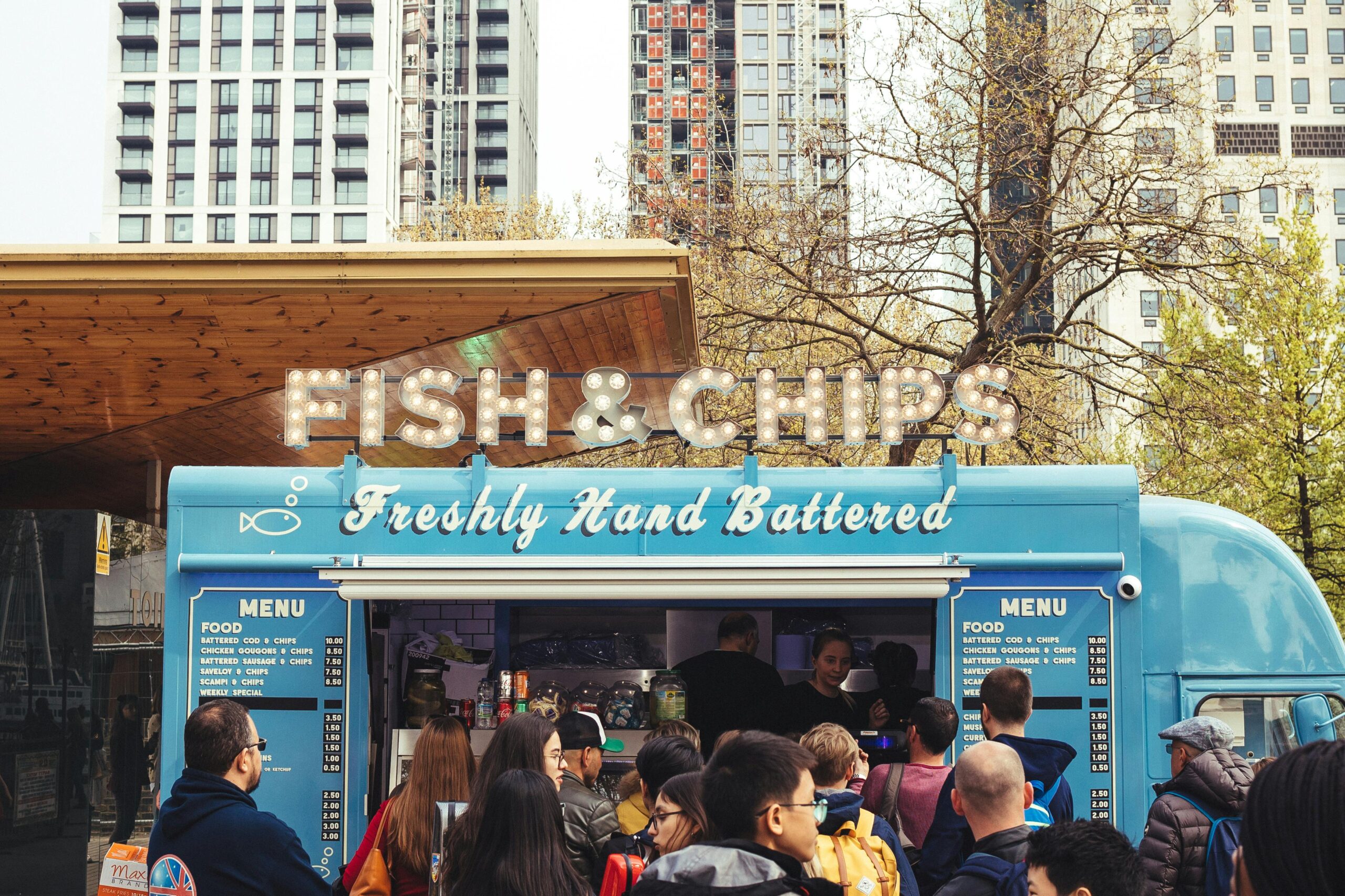 Stock photo of a light blue food truck with dozens of people crowded around waiting in line. Marquee letters above the truck read "Fish & Chips" and cursive white text on the truck reads "Freshly Hand Battered." There is a city in the background with tall buildings.