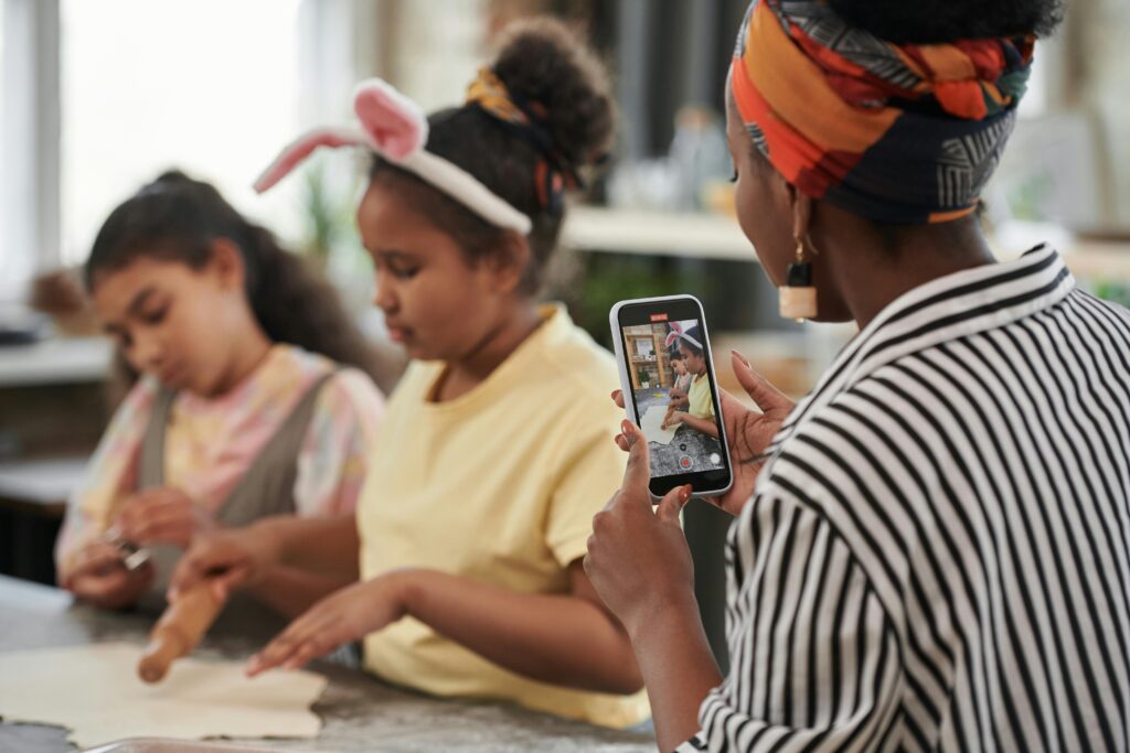 Stock photo of a woman recording two girls rolling out dough on a kitchen counter. The woman is facing the girls and is using her phone camera to videotape them.