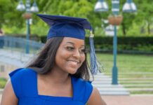Stock photo of a young woman smiling and looking off to the side. She is dressed in a blue graduation gown with a square collar and a matching blue graduation cap with a blue and white tassel.