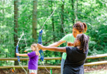Does Your Child Have a Specialized Need? There’s a Camp for That Photo of a woman assisting two children with archery in the woods.