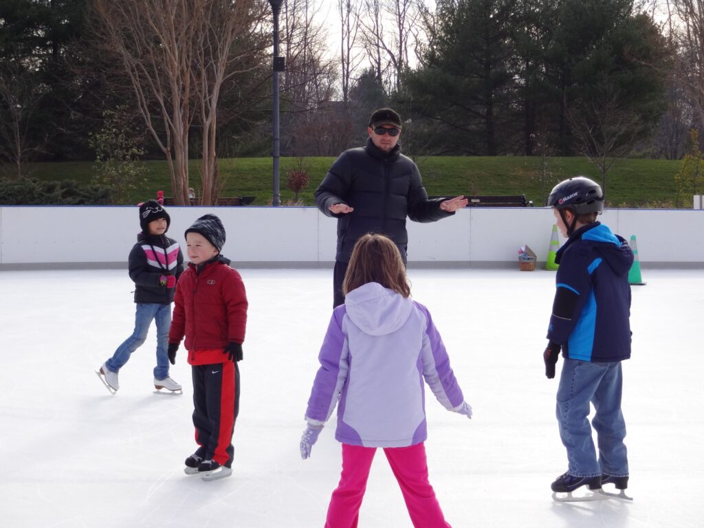 Photo of a man surrounded by four children on the ice at an outdoor ice rink. Everyone is dressed in winter coats and ice skates.