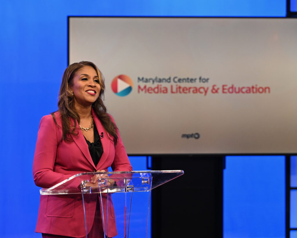 Photo of a woman wearing a hot pink blazer speaking behind a clear podium. Behind her is a large screen that reads "Maryland Center for Media Literacy & Education."