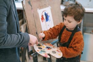 Stock photo of a red-haired toddler holding a paint palette with one hand and a paintbrush in the other. He is standing in front of an easel painting and wearing a black smock.