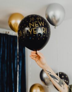 Stock photo of a person holding a black balloon with gold text that reads "Happy New Year!" There are two more balloons in the background, one gold and one silver.