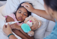 Stock photo of a young girl lying in bed with a teddy bear. An adult is feeling the child's forehead and has a thermometer in the other hand.