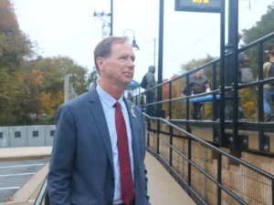 Photo of a man in a suit and red tie standing outside, looking off into the distance.