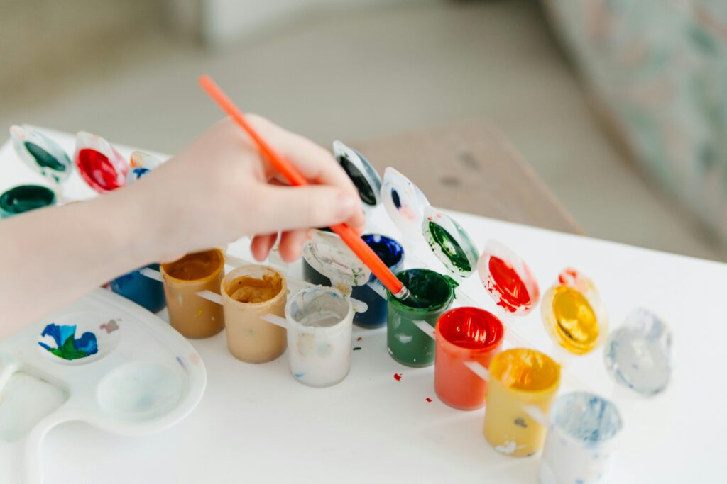 Stock photo of a child's hand holding an orange plastic paintbrush and dipping it in green acrylic paint on a white table.
