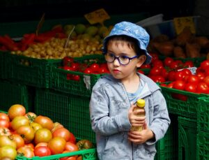 Stock photo of a young boy with round glasses and a blue hat holding a jar of honey at an outdoor fruit stand. He is standing between green crates of red tomatoes.