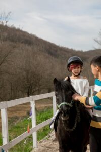 Stock photo of a young girl with short pink hair riding a black pony with the help of an instructor. She is wearing a helmet and is inside a fenced-in area.