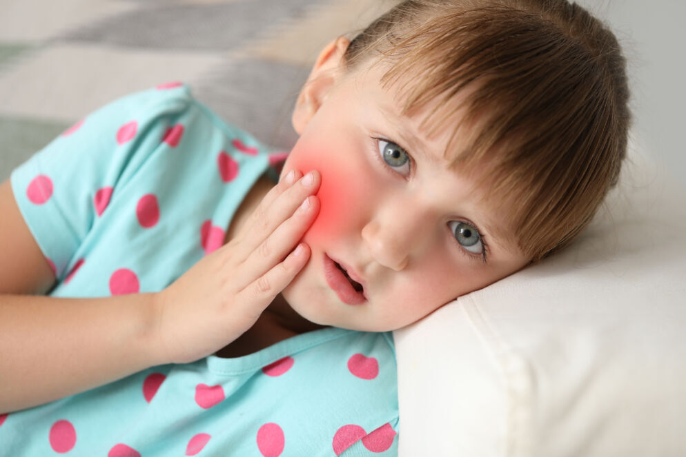 Stock photo of a young girl suffering from a toothache at home. She is lying on a white pillow holding her inflamed cheek in pain.