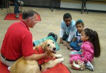 Photo of a man with an arm around a Golden Retriever as three kids listen to the man read a story on a library floor.