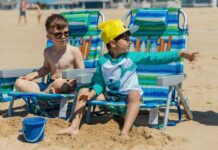 Stock photo of two young boys sitting in beach chairs in the sand. One is wearing sunglasses and swim trunks and the other is wearing a yellow cap, teal swim shirt with sharks on it and swim trunks.