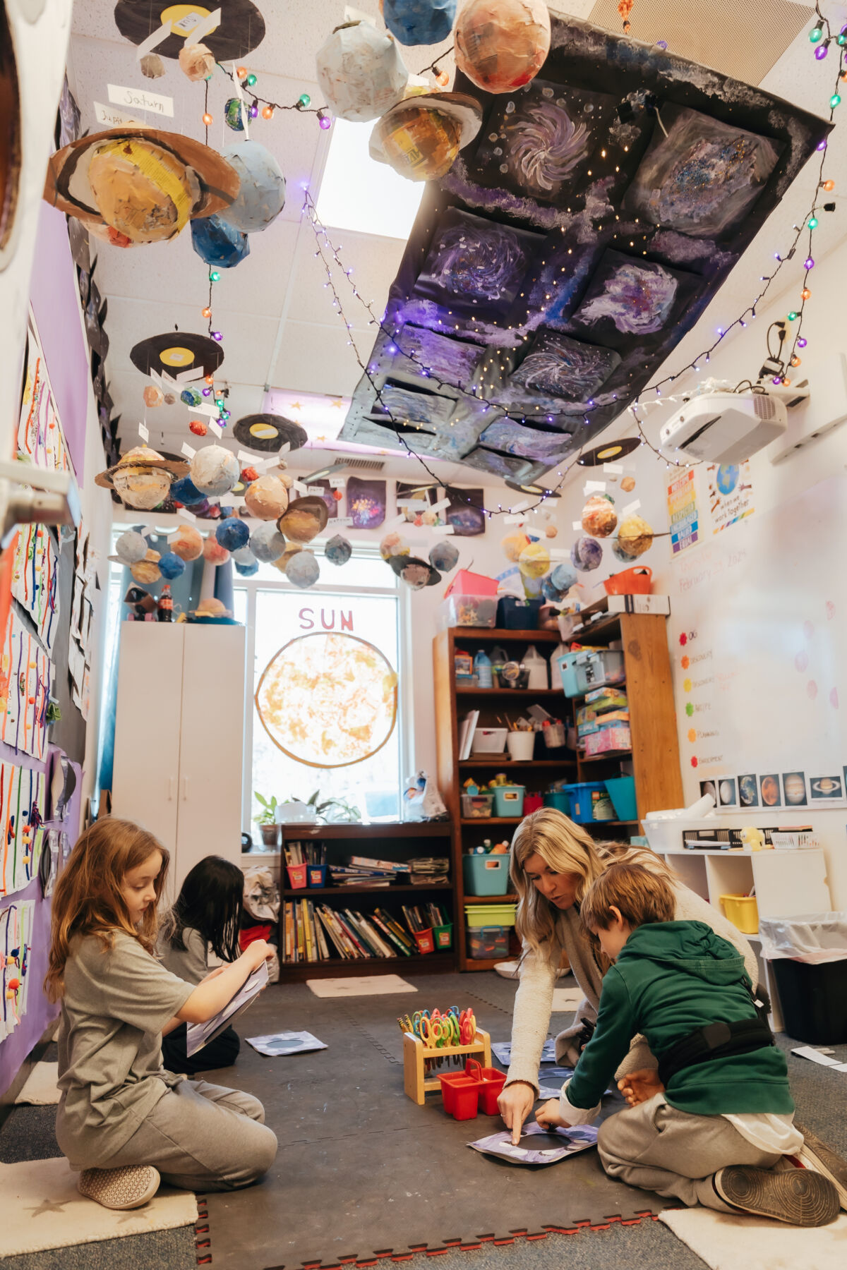 Photo of a classroom with three-dimensional planets hanging from the ceiling. Kids read on the carpet.