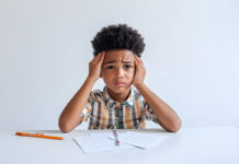 Parents Can Help Support Kids’ Mental Health Throughout the School Year Stressed young boy sits at a desk with papers, holding his head.