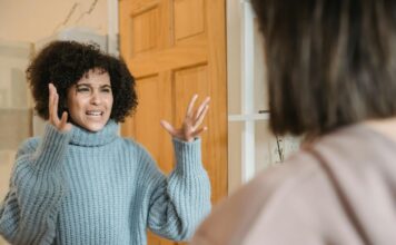 Two women stand by a doorway, one of them shown to be gesturing emphatically with her hands and an expression of frustration.